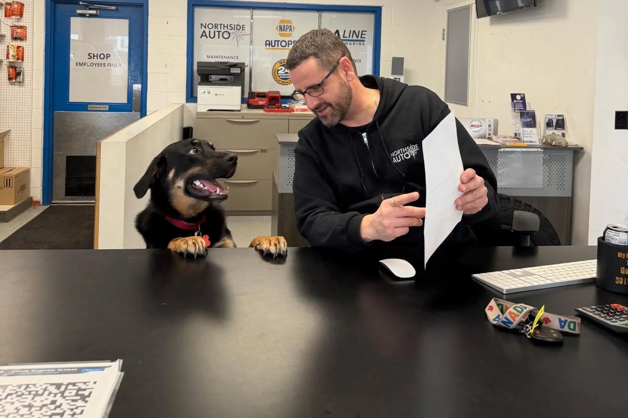 A man behind the shop's front counter holding a sheet of paper, smiling at a dog with its front paws up on the counter